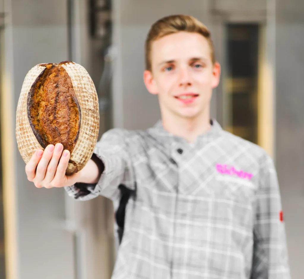 Ein Bäcker in einer karierten Uniform hält einen Laib Brot hoch. Am unteren Bildrand ist der rosa Schriftzug „Jakobs Dinkelkruste“ zu sehen.