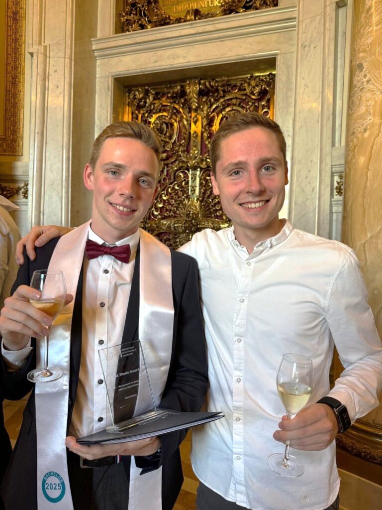 Zwei junge Männer lächeln und posieren bei einer feierlichen Veranstaltung mit Champagnergläsern in der Hand. Der eine trägt einen Anzug, eine Fliege und eine Schärpe mit der Aufschrift „2025“ und hält eine Plakette in der Hand; der andere trägt ein weißes Hemd. Im Hintergrund sind verzierte goldene Türen zu sehen.