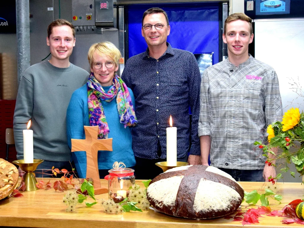 Vier Menschen stehen lächelnd hinter einem Holztisch, auf dem Kerzen, ein Kreuz, Brot und herbstliche Dekorationen stehen. Die Szene scheint in einem Innenraum zu spielen, möglicherweise in einer Küche oder einer Bäckerei, da im Hintergrund Backutensilien zu sehen sind.