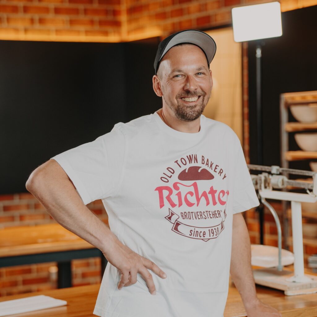 Ein lächelnder Mann in einem weißen T-Shirt und einer Schürze steht in der Küche einer Bäckerei, im Hintergrund sind Holztische, eine Waage und Backsteinwände zu sehen.