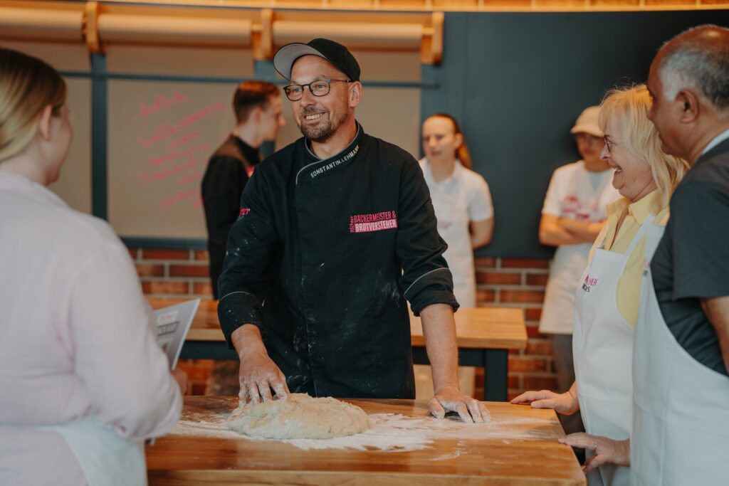 Ein Mann in einer schwarzen Kochuniform knetet Teig auf einem Holztisch und lächelt die Menschen in Schürzen an, die um ihn herumstehen. Die Kulisse scheint eine Bäckerei oder ein Kochkurs zu sein, im Hintergrund sind Backsteinwände zu sehen.