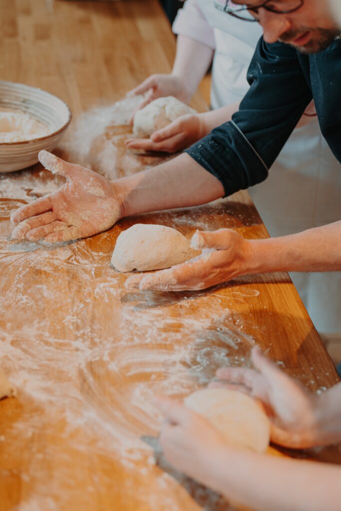 Drei Personen kneten und formen Teig auf einer bemehlten Holzplatte und backen gemeinsam Brot. Ihre Hände sind mit Mehl bedeckt, und in der Nähe stehen Schüsseln.