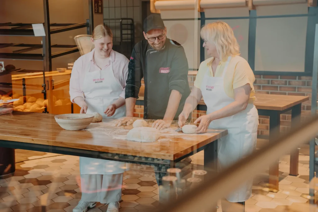 Drei Personen in Schürzen sitzen lächelnd an einem Holztisch in einer Bäckerei und formen gemeinsam Teig, während im Hintergrund Brotlaibe und Backutensilien zu sehen sind.