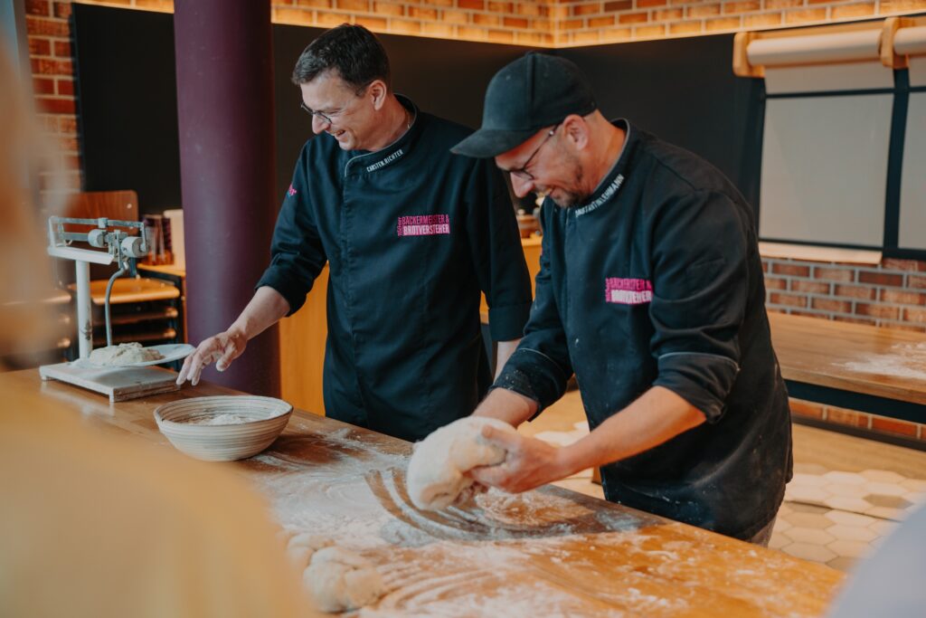Zwei Bäcker in schwarzen Uniformen kneten auf einem bemehlten Holztisch in einer Bäckerei Teig, lächeln dabei und arbeiten zusammen. Backutensilien und eine Schüssel sind zu sehen, im Hintergrund sind Backsteinwände zu erkennen.