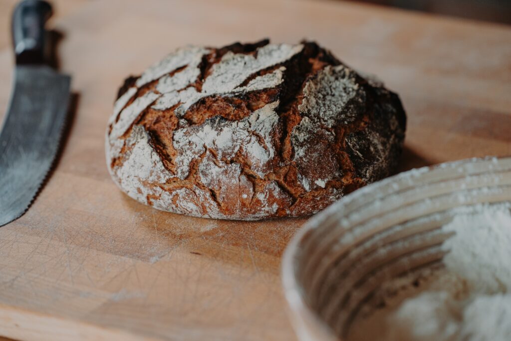 Ein rustikaler Brotlaib liegt auf einer Holzunterlage neben einem gezackten Messer und einem bemehlten Korb, was an eine Szene beim Backen erinnert.