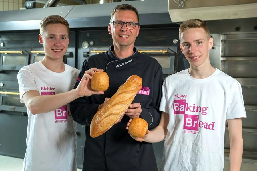 Drei lächelnde Männer stehen in einer Bäckerei und halten frisches Brot in der Hand; der Mann in der Mitte hält ein Baguette, während die beiden Männer an den Seiten Brötchen halten. Die beiden Männer an den Seiten tragen weiße "Baking Bread"-T-Shirts.