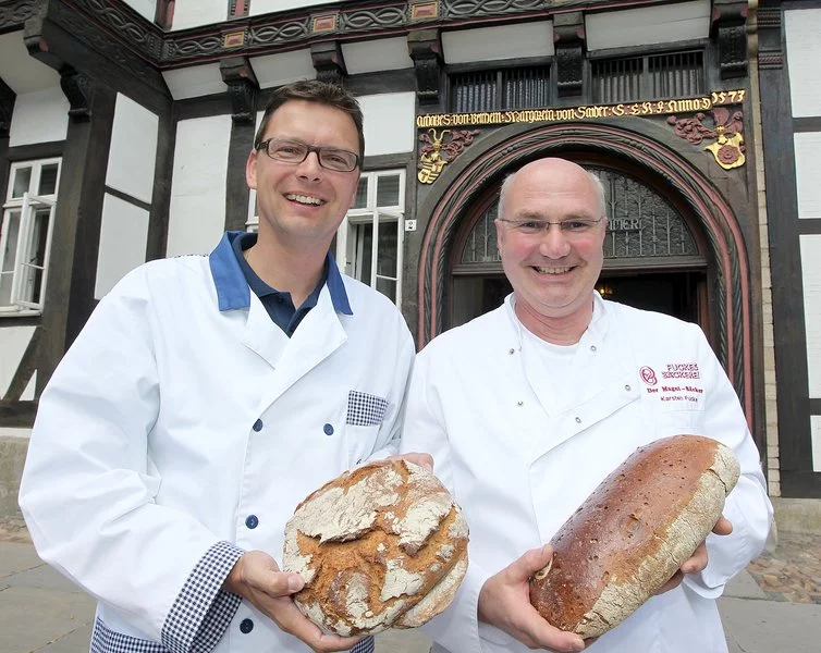 Zwei lächelnde Männer in weißen Kochmänteln stehen im Freien vor einem historischen Gebäude, halten jeweils einen großen runden Laib Brot in der Hand und schauen in die Kamera.