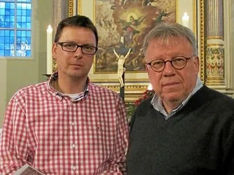 Zwei Männer stehen in einer Kirche, der eine in einem rotkarierten Hemd und mit Brille, der andere in einem dunklen Pullover und mit einer runden Brille. Hinter ihnen befinden sich religiöse Gemälde, ein Kruzifix, Kerzen und kunstvolle Verzierungen.