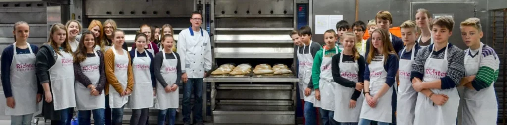Eine Gruppe junger Leute und ein Bäcker in Schürzen stehen in einer Bäckereiküche, versammelt um ein Tablett mit frisch gebackenem Brot vor einem Industrieofen.