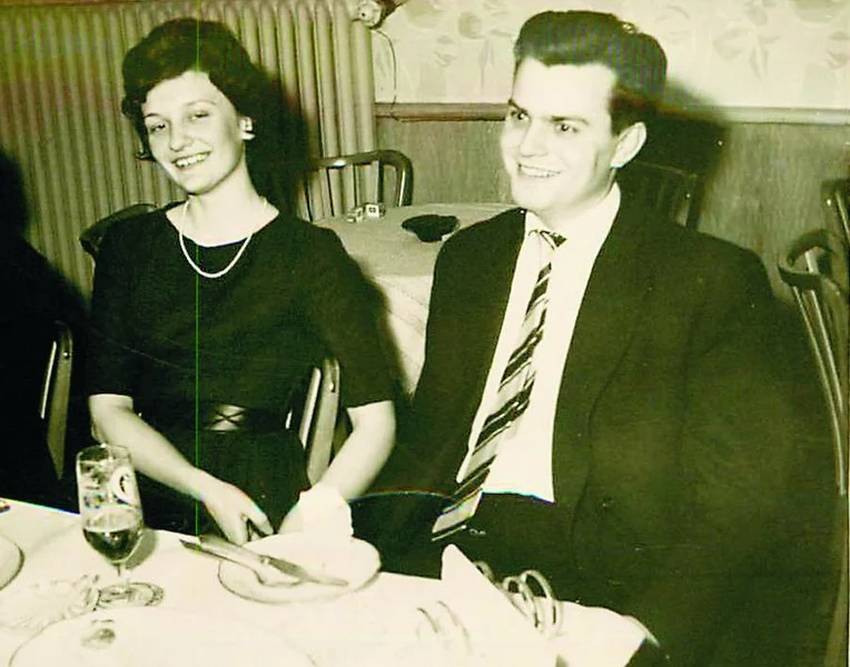 Ein lächelndes, förmlich gekleidetes junges Paar sitzt zusammen an einem für eine Mahlzeit gedeckten Tisch, mit Getränken und Tellern vor ihnen, in einer scheinbar klassischen Restaurant- oder Essensumgebung.
