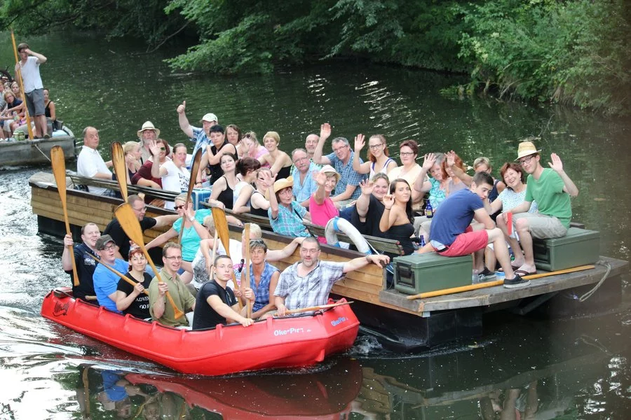 Eine große Gruppe von Menschen, einige in einem roten Kanu und andere auf einem Holzboot, lächeln und winken in die Kamera, während sie auf einem ruhigen, von Grün umgebenen Fluss treiben.