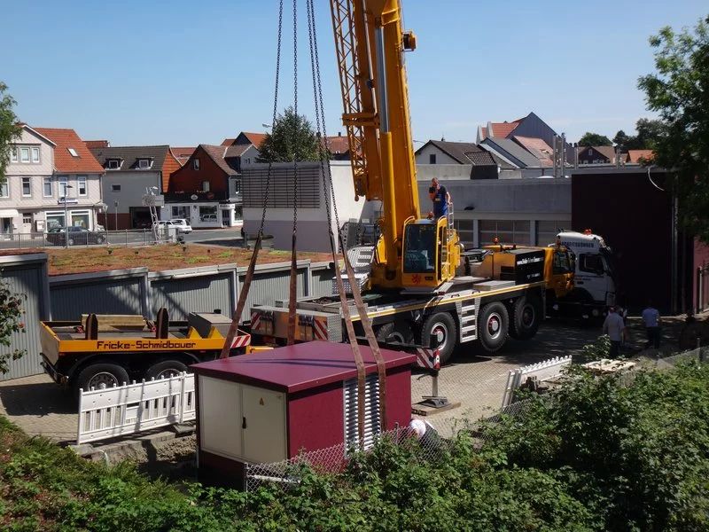 Ein großer gelber Kran hebt eine weiße modulare Bauplatte über ein eingezäuntes Gelände, wobei Arbeiter den Vorgang anleiten. In der Nähe sind im Hintergrund ein gelber Lastwagen und Häuser mit roten Dächern zu sehen.