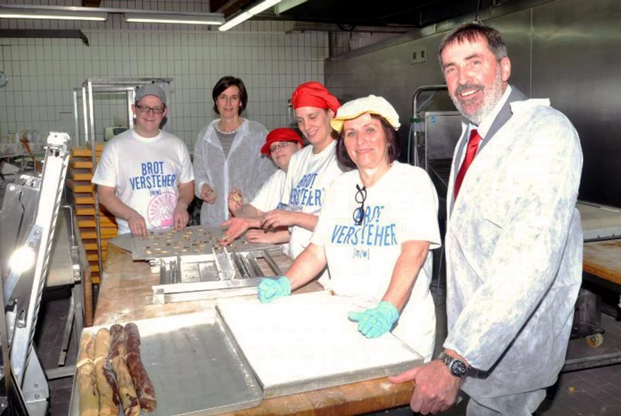 Fünf Bäckereiarbeiter in weißen Kleidern und Hüten bereiten Teig auf einem Tisch vor, während ein Mann im Anzug neben ihnen in einer Bäckereiküche lächelt. Im Hintergrund sind Brotlaibe und Tabletts zu sehen.
