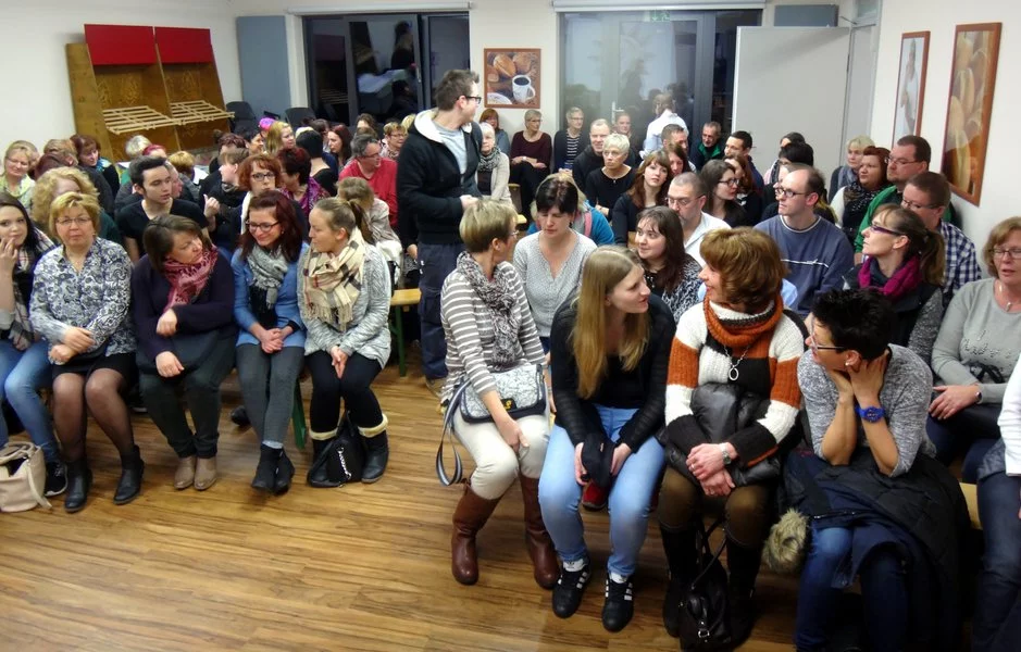 Eine große Gruppe Erwachsener sitzt dicht an dicht auf Stühlen in einem hell erleuchteten Raum mit Holzfußboden, unterhält sich und wartet auf den Beginn einer Veranstaltung. Einige schauen nach vorne, während andere sich in kleinen Gruppen unterhalten.
