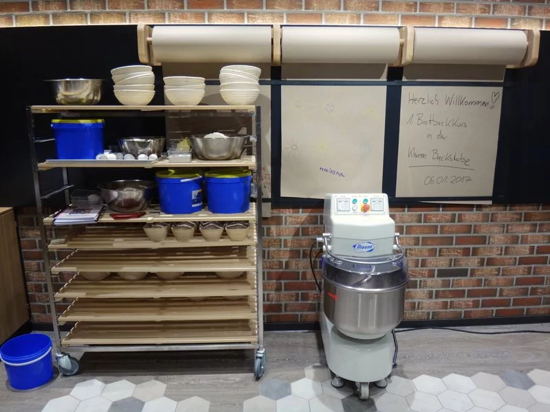 Ein Metallregal mit Schüsseln, Behältern und Tabletts steht neben einem großen Teigmischer. An der Wand darüber hängen braune Papierrollen, und in der Nähe ist ein handgeschriebenes Willkommensschild in deutscher Sprache angebracht. Der Boden ist mit sechseckigen Fliesen belegt.