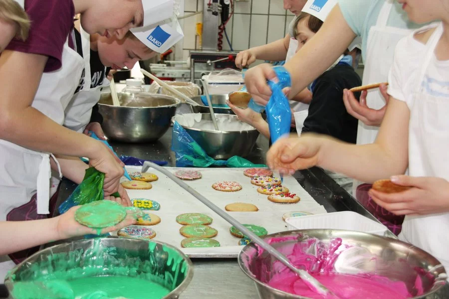 Kinder in Schürzen verzieren Kekse mit buntem Zuckerguss und Streuseln an einem Küchentisch. Schüsseln mit grünem und rosa Zuckerguss, Spritzbeutel und verzierte Kekse sind in einer geschäftigen, kreativen Atmosphäre zu sehen.