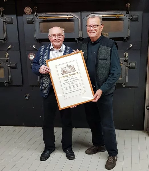 Zwei ältere Männer stehen in einem Haus, lächeln und halten eine gerahmte Urkunde in der Hand. Hinter ihnen stehen große Industrieöfen mit Metalltüren und Drehknöpfen, die an eine Bäckerei oder eine Großküche erinnern.