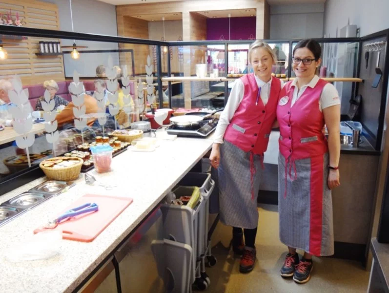 Zwei Frauen in passenden rosafarbenen Westen und karierten Schürzen stehen lächelnd hinter einem Tresen in einem modernen Café oder einer Bäckerei, wo Lebensmittel und Vorräte sichtbar sind. Im Hintergrund sitzen Menschen und essen.