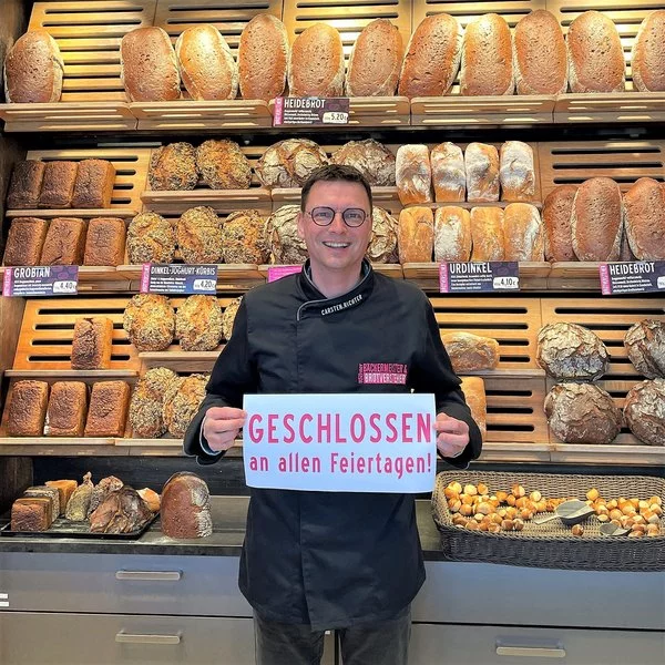 Ein lächelnder Bäcker in schwarzer Uniform steht vor Regalen, die mit verschiedenen Brotsorten gefüllt sind, und hält ein Schild mit der Aufschrift GESCHLOSSEN an allen Feiertagen! (Geschlossen an allen Feiertagen! auf Deutsch).