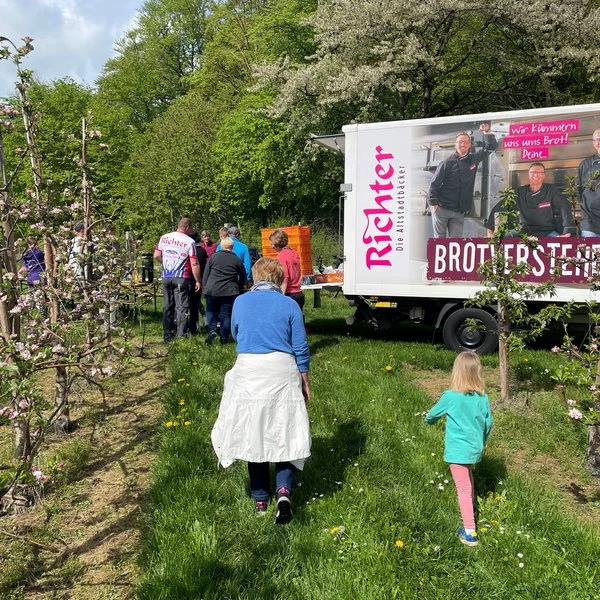 Menschen gehen durch einen grasbewachsenen Obstgarten auf einen weißen Imbisswagen mit rotem Richter-Branding zu. Erwachsene und ein Kind sind zu sehen, und blühende Bäume umgeben die Szene an einem sonnigen Tag.
