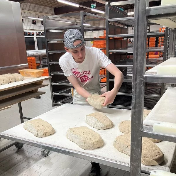 Ein junger Mann mit Mütze und "Baking Bread"-T-Shirt formt auf einem bemehlten Tisch in einer Bäckerei, umgeben von leeren Metallgestellen und Tabletts, Teigbrote.