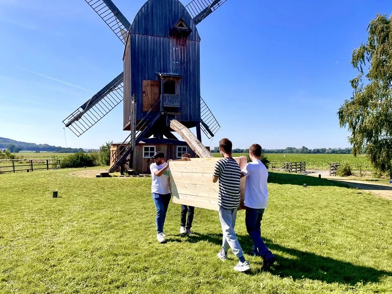 Vier Personen tragen an einem sonnigen Tag ein großes Holzbrett über eine grasbewachsene Wiese zu einer Windmühle mit Holzflügeln. Die Windmühle steht im Hintergrund, umgeben von grünen Feldern und einem klaren blauen Himmel.