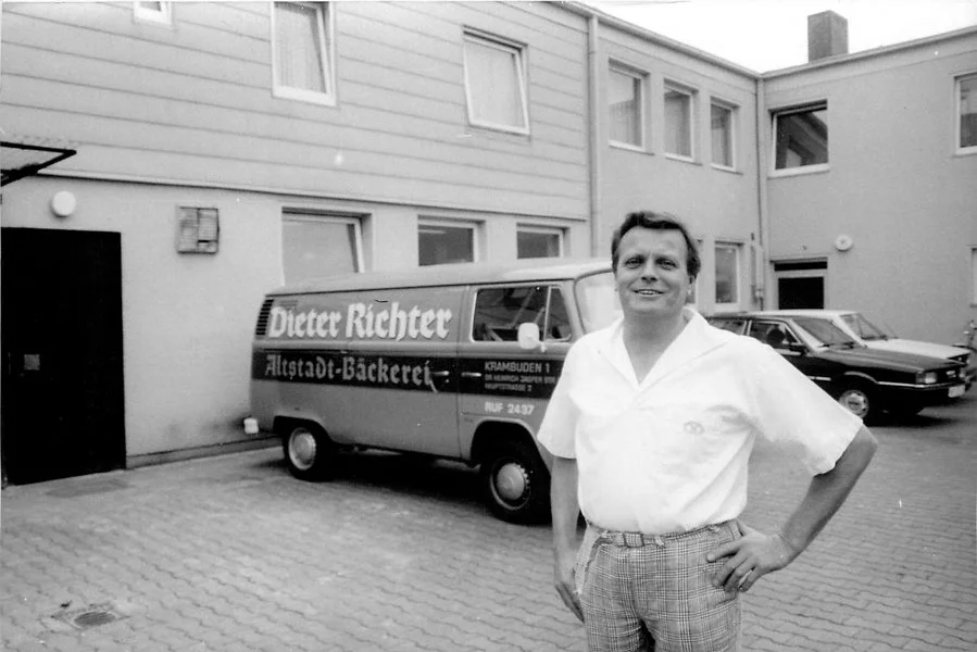 Ein lächelnder Mann im weißen Hemd steht in einem gepflasterten Innenhof und hat die Hand in die Hüfte gestemmt. Hinter ihm stehen ein Lieferwagen mit der Aufschrift Dieter Richter Altstadt-Bäckerei und ein Gebäude mit mehreren Fenstern.