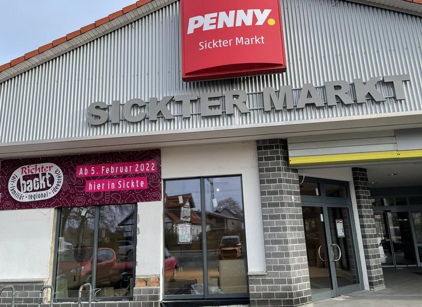 Außenansicht eines Gebäudes mit den Schildern PENNY. Sickter Markt und SICKTER MARKT. Ein Banner wirbt für Richter Dacht ab 5. Februar 2022. Es gibt Fenster und eine gläserne Eingangstür.