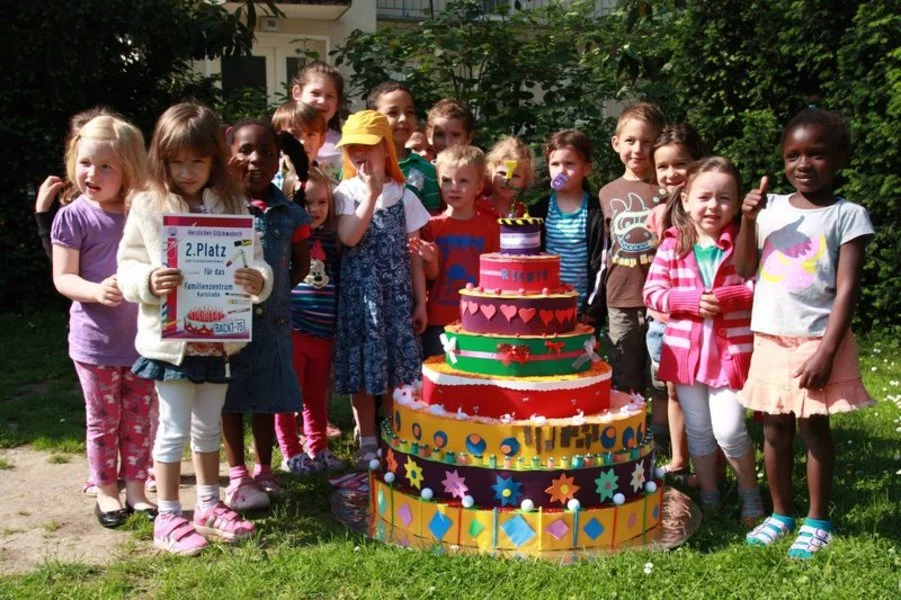Eine Gruppe junger Kinder steht im Freien um eine große, farbenfrohe, gestufte Kuchendekoration herum. Einige Kinder lächeln und posieren, während eines eine Urkunde in der Hand hält. Im Hintergrund sind grüne Sträucher und Gras zu sehen.