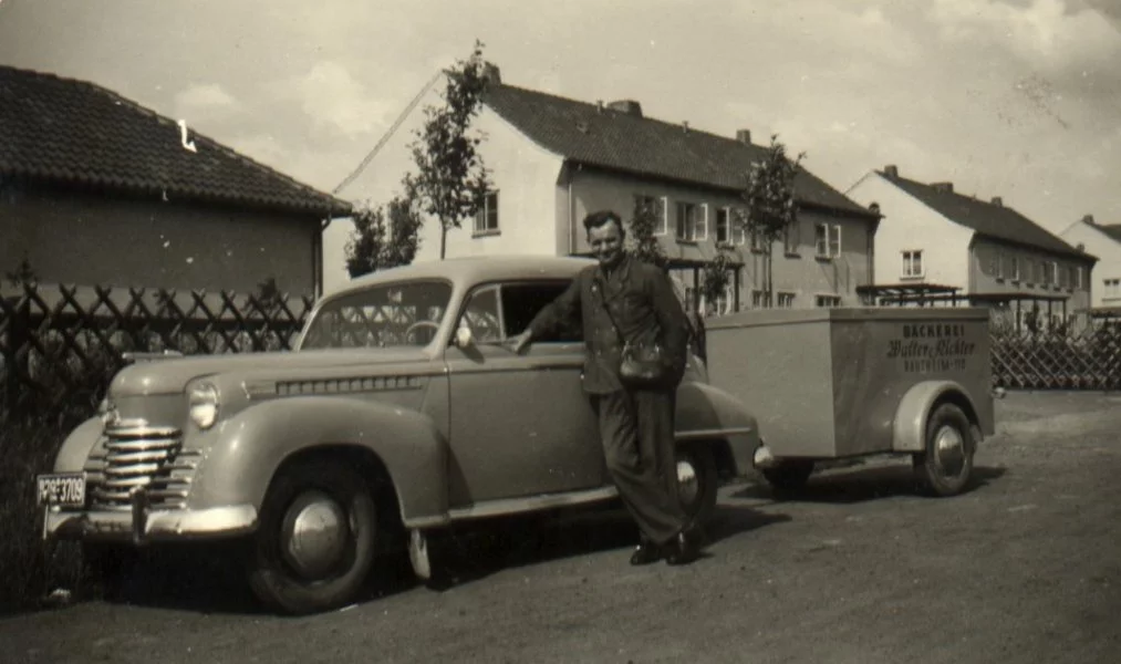 Ein Mann in einer Militäruniform lehnt an einem Oldtimer mit Anhänger, der in einer Wohnstraße mit Häusern und kleinen Bäumen im Hintergrund geparkt ist.