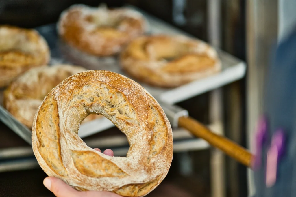 Eine Person hält einen runden, rustikalen Brotlaib mit einem Loch in der Mitte in der Hand. Im Hintergrund liegen weitere ähnliche Brotlaibe auf einem Backblech, frisch aus dem Ofen.